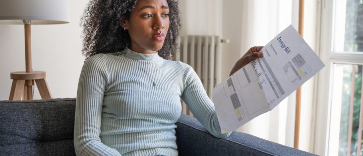 A woman sitting on a sofa holding an energy bill and looking worried